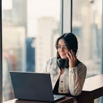 Young woman in a modern office using a laptop and headset, gazing out the window at the cityscape.